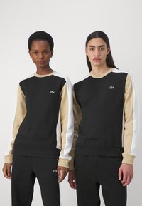 Felpe nere e beige con accenti bianchi, dotate di collo a giro e logo ricamato. Tessuto morbido, vestibilità casual, abbinate a pantaloni neri.