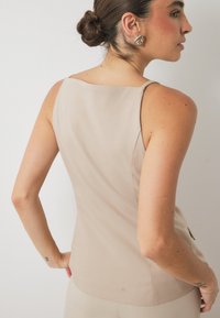 Bež brezrokavnik iz gladkega materiala s poenostavljeno obliko, ki ima kvadratno zadnjo izreznico in prilegajočo se silhueto.