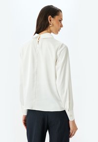 Weiße Bluse mit einem Schlüsselspalt auf der Rückseite, langen Ärmeln und Knopfmanschetten. Glatter Stoff, taillierte Passform und subtiler Glanz. Kombiniert mit gestreiften Hosen.