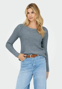 Femme blonde portant un pull en maille gris rentré dans un jean taille haute bleu clair avec une ceinture marron, posant avec une main dans la poche.