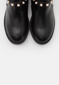 Botas de tobillo de cuero negro con punteras redondas, que presentan una correa con detalles dorados adornada con perlas sintéticas y suelas de goma texturizadas.