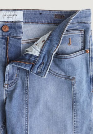 Vaqueros de corte ajustado en azul claro con cremallera parcialmente abierta, botón de cobre, pequeño bolsillo delantero con bordado en forma de "J" y etiqueta blanca de la marca "Jeckerson".