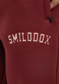 Bordeaux joggingbroek met geborduurd "SMILODOX" logo in contrasterend crème. Zachte textuur met ribgebreid materiaal en ritszakdetails.