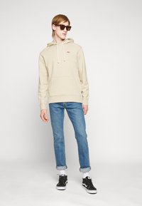 Beige hettegenser med lommer foran og snorer, kombinert med blå jeans og svarte joggesko. Modell som har på seg mørke solbriller.