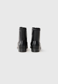 Un par de botas de cuero negro hasta el tobillo con tacones bajos, vistas desde atrás sobre un fondo blanco liso.