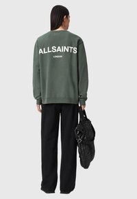 Oliwkowy sweatshirt z napisem "ALLSAINTS LONDON" w białym kolorze na plecach, zestawiony z luźnymi czarnymi spodniami i czarną torbą.