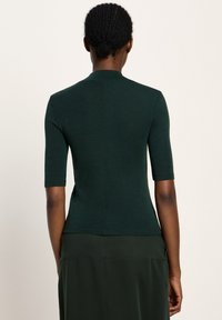 Woman with braided hair bun wearing a dark green short-sleeve fitted top and matching skirt, standing facing away against a plain background.