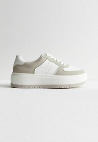 Zapatilla con parte superior de cuero blanco y detalles en ante gris, con perforaciones y una suela blanca gruesa con agarre texturizado.