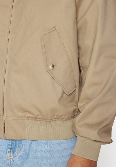 Veste bomber beige avec fermeture éclair, poignets côtelés et une poche zippée sur le côté gauche avec un détail de bouton circulaire.