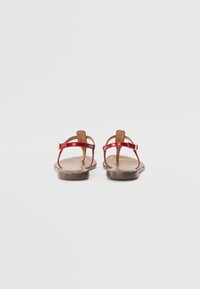 Red flat sandals with thin straps and a thong design. Textured brown footbed with a geometric pattern. Gold buckle detailing on straps.