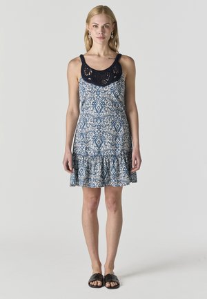 Szőke nő áll, kék-fehér mintás ujjatlan ruhát visel fekete horgolt nyakkivágással és fekete pántos szandállal.