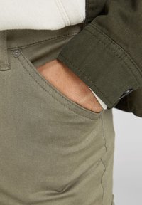 Eine Hand steckt in der vorderen Tasche einer olivgrünen Hose, dazu wird eine dunkelgrüne Jacke getragen, deren Ärmel teilweise über einem hellen Pullover sichtbar ist.