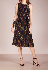 Schwarzes Blumenkleid mit gerafftem Ausschnitt, ärmellosem Design, Rüschung am Saum und einem passenden Gürtel; kombiniert mit schwarzen Riemchenheels und einer Handtasche.