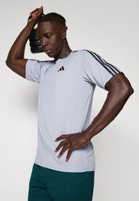 Lichtgrijs sportshirt met korte mouwen en zwarte strepen op de schouders en logo. Het model houdt een zwarte kettlebell vast, wat een gespierd figuur toont.
