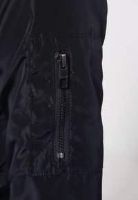 Černá nylonová bunda s kapsou na zip, hladkou texturou a jemným leskem. Zip má stříbrný přívěsek a značkové písmo.