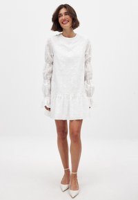 Balta suknelė su siuvinėtu gėlių raštu, ilgomis pūstomis rankovėmis, raukšlėtu apdailu ir apvaliu kaklu. Dėvima su smailiais baltais bateliais.