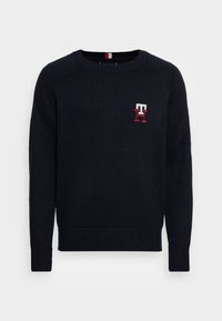 Marineblauwe gebreide trui met geribbelde manchetten en zoom. Voorzien van een klein geborduurd logo in wit en rood op de linkerborst. Rondhals ontwerp.