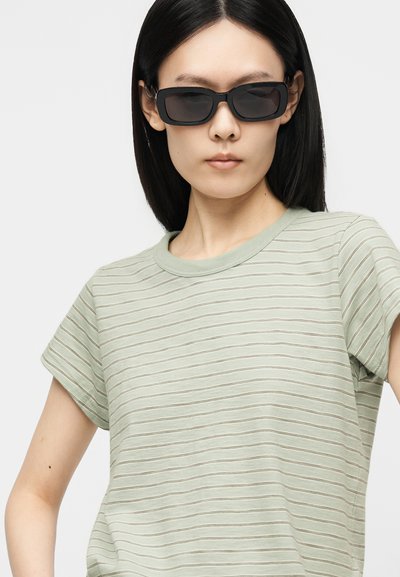 Γυναίκα με ίσια μαύρα μαλλιά φοράει ορθογώνια μαύρα γυαλιά ηλίου και μια ανοιχτοπράσινη ριγέ κοντομάνικη μπλούζα σε ένα απλό φόντο.