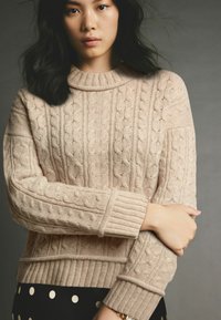 Beige gestrickter Pullover mit Zopfmuster, hohem Kragen und gerippten Bündchen. Auffällige Textur und Dicke. Kombiniert mit einem schwarzen Rock mit Punkten.