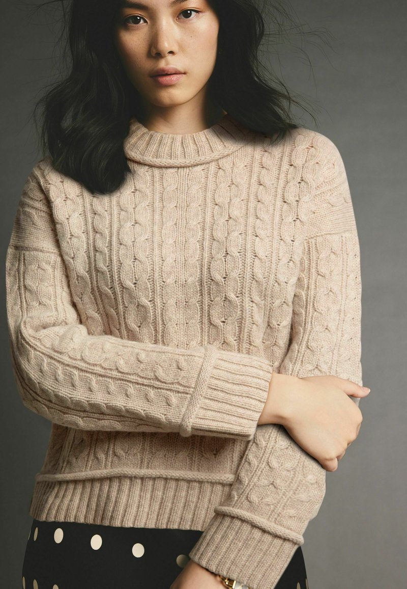 Beige gestrickter Pullover mit Zopfmuster, hohem Kragen und gerippten Bündchen. Auffällige Textur und Dicke. Kombiniert mit einem schwarzen Rock mit Punkten.