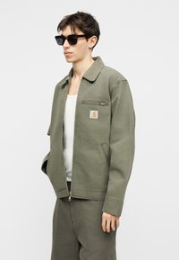 Νέος άνδρας που φοράει γυαλιά ηλίου με σκελετό από χελώνα, πράσινο μπουφάν Carhartt με φερμουάρ πάνω από λευκό πουκάμισο και ταιριαστό πράσινο παντελόνι, όρθιος με τα χέρια στις τσέπες.