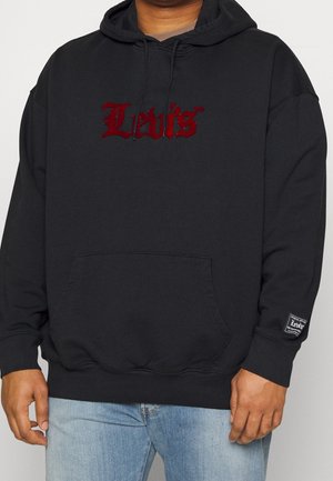 Mężczyzna w czarnej bluzie z kapturem z czerwonym logo Levi’s na piersi i naszywką Levi’s na lewej rękawie, w połączeniu z jasnoniebieskimi dżinsami.