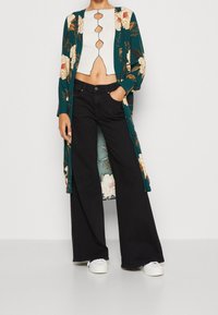 Dlouhý květinový kimono v odstínu teal a krémové s hlubokým výstřihem, spojený s černými širokými kalhotami a bílými teniskami. Obsahuje květinový vzor.