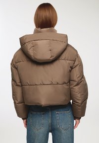 Brązowa krótka kurtka puchowa z kapturem, z pikowanymi sekcjami i gładkim wykończeniem. Nosi ją osoba w niebieskich dżinsach.