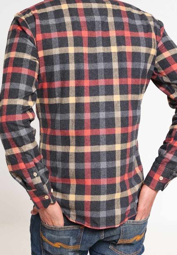Uomo che indossa una camicia a maniche lunghe scozzese rossa, beige e nera, con le mani nelle tasche posteriori di un paio di jeans blu.