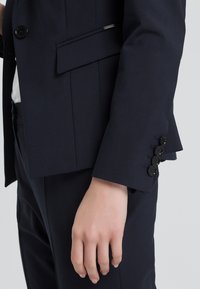 Navy tailored blazer featuring structured lapels, a single button closure, and four black buttons on the cuff. Smooth fabric texture.