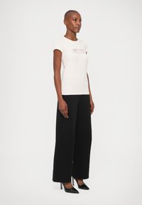 Tricou alb ajustat cu logo-ul "ARMANI EXCHANGE", combinat cu pantaloni negri cu picior larg și tocuri negre ascuțite. Material neted, design simplu.