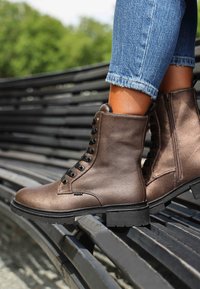 Botas de tobillo de cuero marrón con cordones negros en la parte frontal, superficie texturizada, suela gruesa negra y detalle de cremallera lateral, situadas sobre un banco.