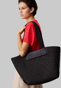 Młoda kobieta w czerwonej koszuli trzyma dużą czarną wiklinową torbę na ramię z szerokimi ramiączkami, stojąc na szarym tle.