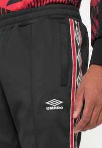 Schwarze Sporthose mit roten und weißen Rauten-Musterstreifen. Mit einem elastischen Bund und einem sichtbaren weißen Umbro-Logo.