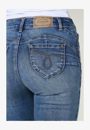 Sinine denimist teksapüksid, mis on lõike poolest mugavad ja millel on pruunid õmblused ning südamekujulised taskud. Nahast brändi silt vöökoha juures.