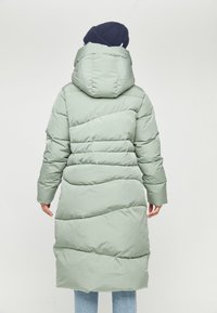 Pitkä muhkea takki vaaleanvihreänä, jossa on huppu, vino ommeltu kaavoitus ja ribatoitu tummansininen pipom. Valmistettu sileästä, eristetyistä materiaaleista.