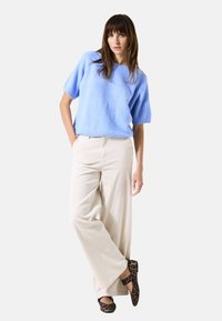 Pull en tricot à manches courtes bleu clair, pantalon beige à jambes larges et chaussures imprimé léopard. Le modèle se tient avec une main dans la poche, devant un fond uni.