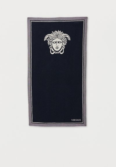 Μαύρη ορθογώνια πετσέτα με γκρι περίγραμμα, που φέρει ένα λευκό σχέδιο κεφαλής Μέδουσας στην κορυφή και το κείμενο «VERSACE» στην κάτω δεξιά γωνία.