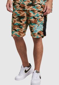 Shorts camouflés dans des nuances de brun, de vert et de bleu, présentant un logo sur la jambe gauche et des accents noirs sur les côtés. Portés avec des baskets blanches.