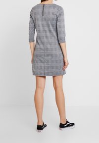 Grå rutig klänning med trekvartslånga ärmar, dragkedjedetalj på ryggen och A-linjeform. Bärs med svarta sneakers. Slät textur och skräddarsydd passform.