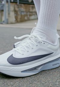Bijela sportska cipela s teksturiranim gornjim dijelom, tamnim Swoosh logom i ZoomX amortizacijom. Uparena s bijelom rebrastom čarapom na asfaltiranoj površini.
