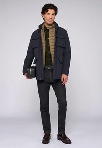 Granatowa parka z wieloma przednimi kieszeniami, nałożona na beżową pikowaną kamizelkę. Zielona koszula pod spodem, ciemne dżinsy i brązowe buty. W ręku czarna torba.