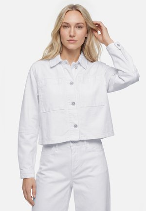 Frau mit langen blonden Haaren, die eine weiße Jacke mit Knöpfen und weiße Hose trägt und mit einer Hand ihr Haar berührt.