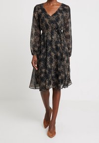 Svart klänning med V-ringning, långa genomskinliga ärmar och ett mönster av guldprickar. Fluffig kjol med insydd midja, bärs med bruna spetsiga klackar.