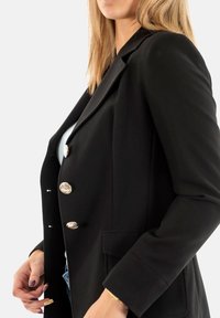 Blazer negro con un corte ajustado, que presenta dos bolsillos laterales y tres botones dorados ornamentados. Tejido suave y solapas estructuradas.