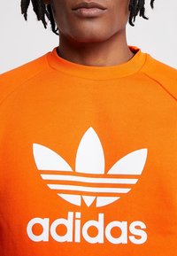 Jasně oranžová mikina s žebrovaným límcem, na které je velké bílé logo Adidas a nad ním tři vodorovné pruhy. Jemná struktura látky.