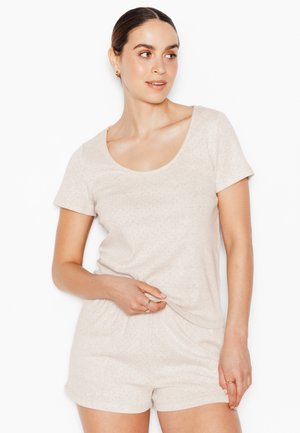 Donna con i capelli tirati indietro che indossa un top beige chiaro a maniche corte e pantaloncini abbinati, guarda a destra mentre si sistema la camicia con la mano.