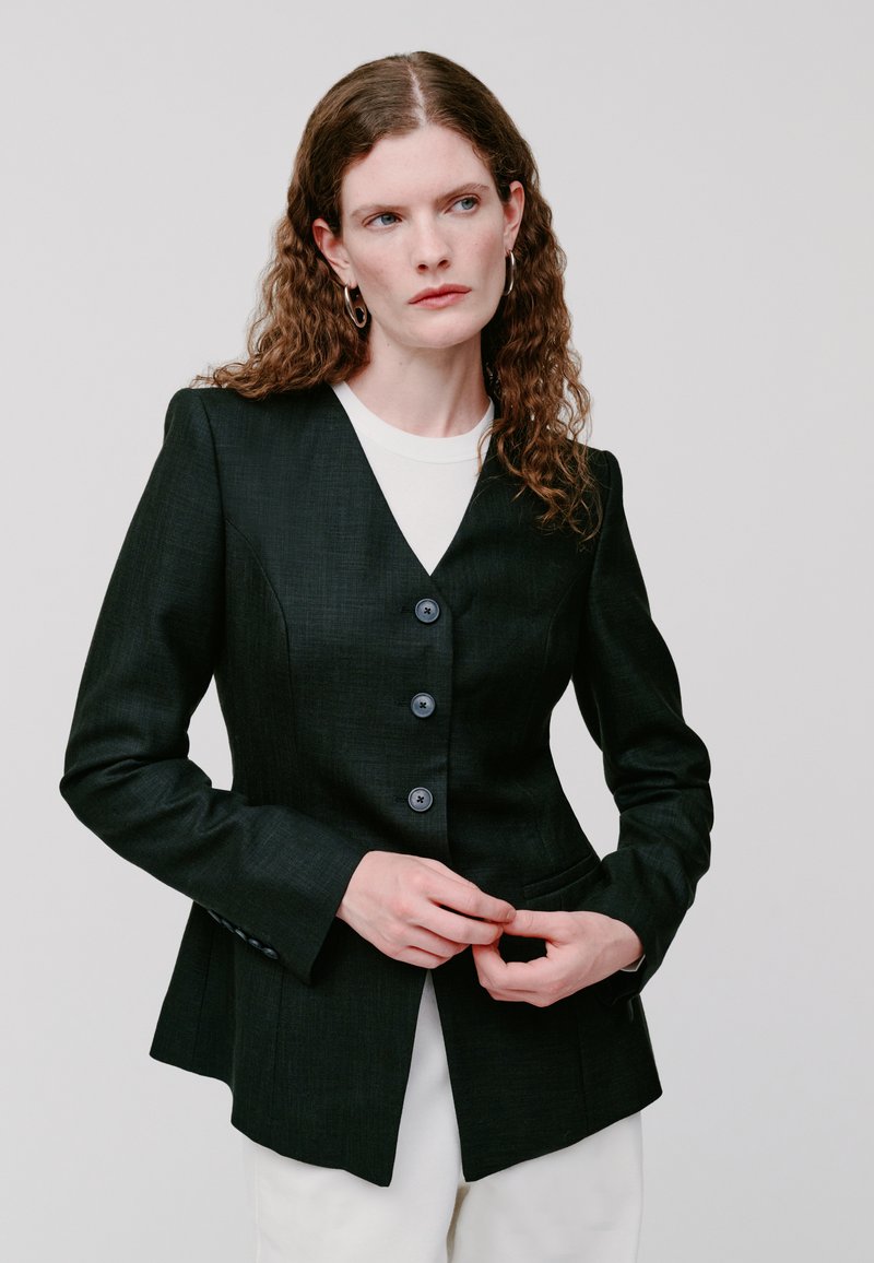 Blazer su misura verde scuro con scollatura a V, quattro bottoni e spalle strutturate. Presenta tasche e tessuto texturizzato.