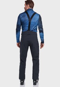 Mann mit schwarzen Skihosen und Trägern sowie einem blauen Langarmoberteil, der mit dem Rücken zu einer einfarbigen hellen Hintergrund steht.