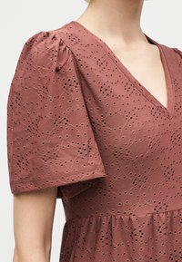 Κοντινό ενός ατόμου που φοράει ένα σκουρόχρωμο πορτοκαλί φόρεμα με τρύπες, φουσκωτά κοντά μανίκια και V-λαιμόκοψη.